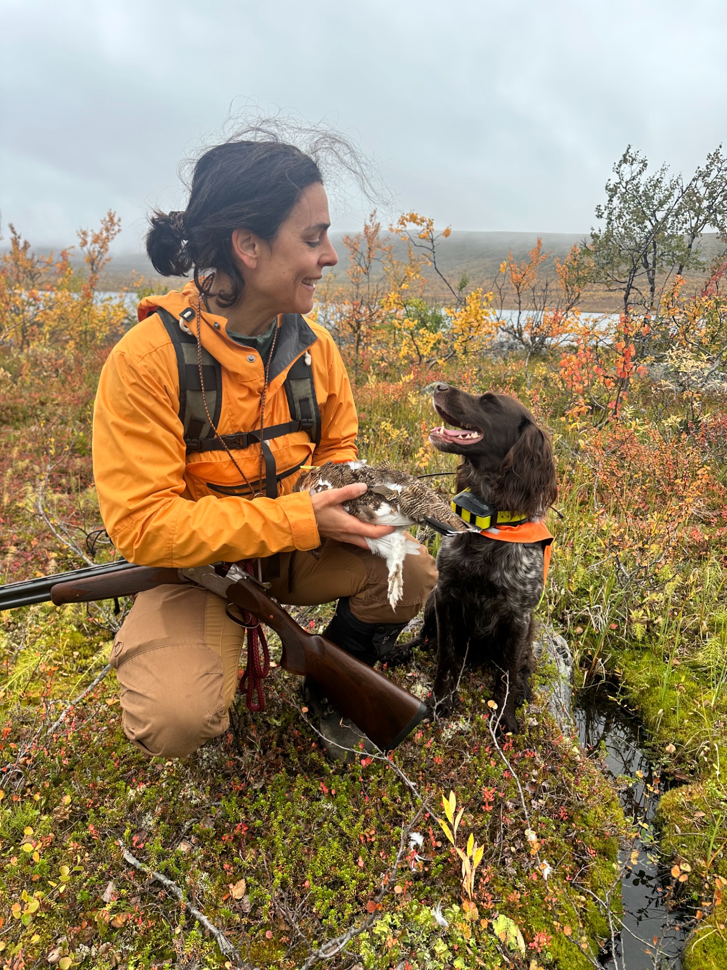 Woman in orange jacket holds a black-and-white dog outdoors, smiling amid autumn foliage and hills.